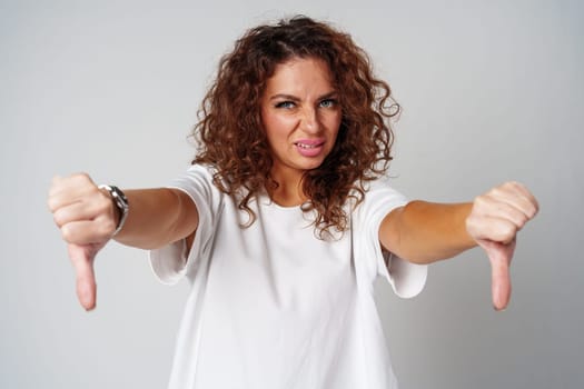 A woman with curly hair shows dislike with thumbs down against a neutral background in a casual setting