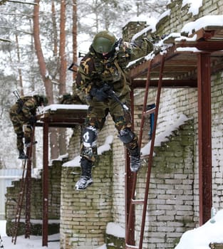 Special forces soldiers climbing a building in combat gear during a military exercise. Armed with rifles, helmets, and protective gear. Brick building with windows in the woods.