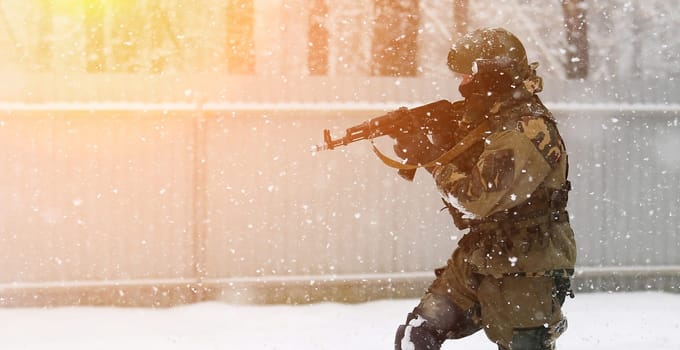 A soldier wearing a mask and winter camouflage uniform is pictured advancing through the snow while holding a gun. The soldier is likely participating in a military operation or training exercise.