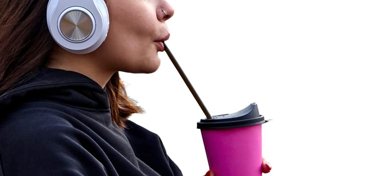 A young woman with headphones takes a sip from a pink coffee cup. She appears relaxed and content, enjoying her beverage in the fresh air.