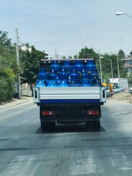 A truck carries blue gas cylinders in an open body along a city face, rear view.