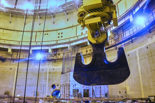 A large, steel claw hangs suspended from a crane inside a vast, concrete structure. The claw appears to be reaching towards a shaft of sunlight that illuminates the room. The structure appears to be under construction and is surrounded by scaffolding and other building materials.