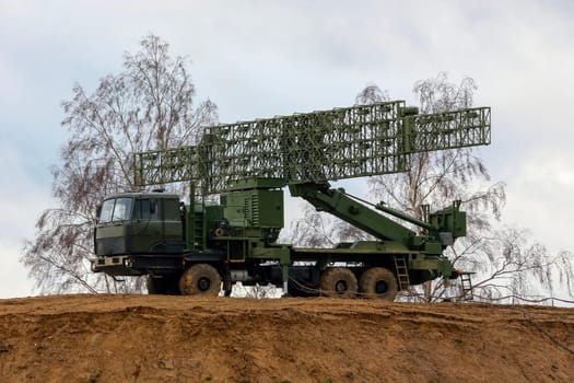 A large, military radar system is mounted on a truck and positioned on a dirt hilltop. The radar dish is extended and pointed upwards, likely scanning for potential threats in the sky.