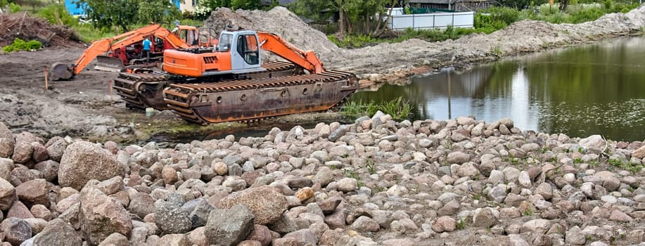 An orange and gray amphibious excavator works on the edge of a pond, placing large rocks to stabilize the shoreline. The excavator's tracks are partially submerged in the water.
