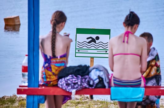 On a bright, sunny day by the tranquil lakeside, two friends sit closely on a red bench, wrapped in vibrant towels and swimwear, engaged in quiet conversation.