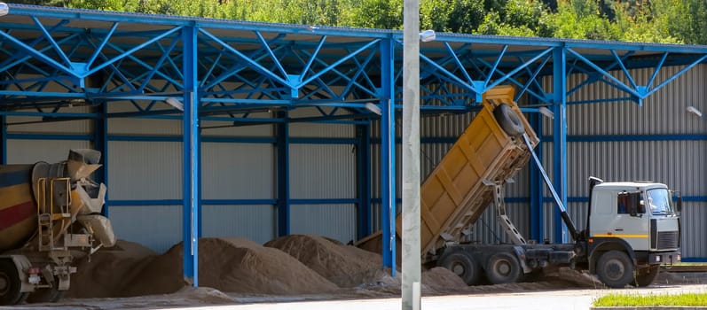 In a bustling construction area, trucks unload their cargoes of sand and gravel under bright blue skies. A large yellow dump truck raises its bed, releasing a cascade of material into a designated space. Nearby, concrete mixers wait patiently, their large compartments filled with a mixture prepared for upcoming projects.