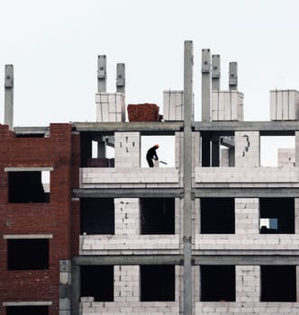 A solitary worker is seen meticulously laying bricks on the scaffolding of a rising multi-story building. Surrounding him are the skeletal remains of the structure, showcasing unfinished concrete and brickwork. Above, gray clouds hang low, hinting at impending rain, while the morning light filters in, casting shadows across the building.