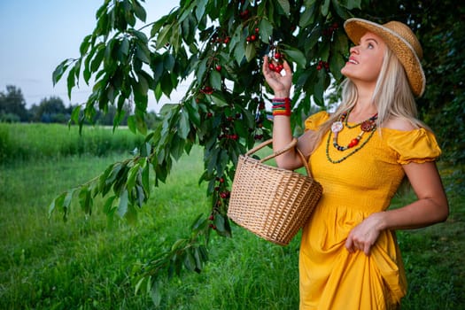 A smiling blonde woman holds ripe red cherries under a cherry tree with heavy fruit-laden branches. She is wearing a mustard-colored dress and stands amidst green leaves on a warm spring day.