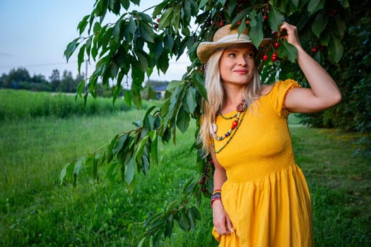 A smiling blonde woman in a mustard dress holds ripe red cherries in her hands while standing by a cherry tree with green leaves. It's a warm, sunny spring day.