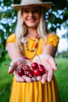 A smiling farmer woman in a mustard-colored dress offers freshly picked red cherries. Her hands reach toward the camera, with a green grassy backdrop in soft focus.