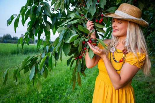 A smiling blonde woman, dressed in a mustard-colored dress, stands by a cherry tree with low-hanging branches full of ripe red cherries. It's a warm spring day in a peaceful orchard.