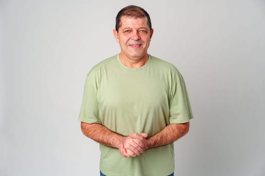 Middle-aged man smiling warmly while standing with hands clasped in front of light gray background close up