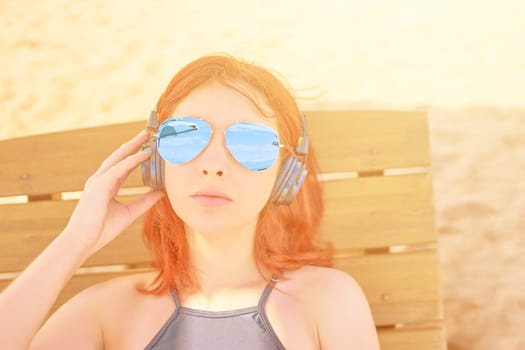 Pretty woman listening to music on the beach. Redhead sexy woman listening melody with earphones while relaxing on beach lounger near sea.