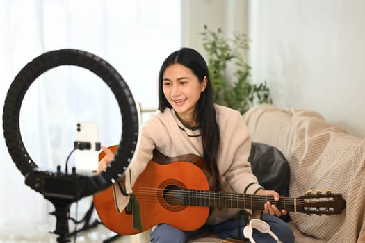 Pretty young artist teaching guitar online recording her lesson with smartphone at home.
