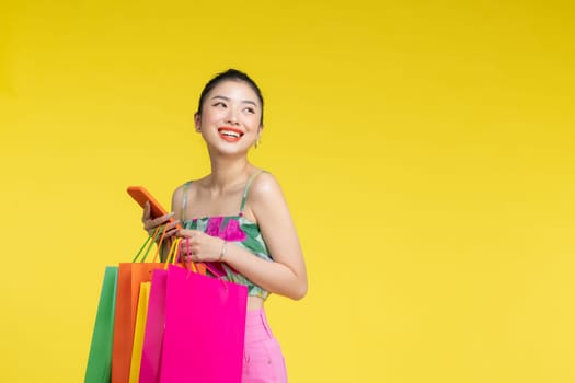 Young energetic Asian woman holding mobile phone with shopping bags on yellow background