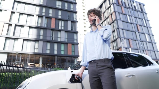 Suit-clad businessman with progressive ambition leaning on his electric vehicle while standing on a charging station with a power cable plug and a renewable energy-powered electric vehicle.