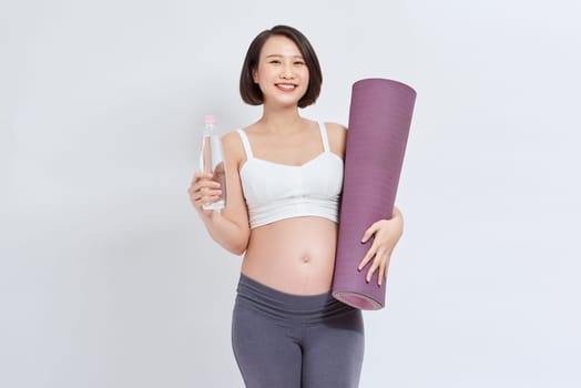 Young pregnant woman holding yoga mat and bottle of water on white
