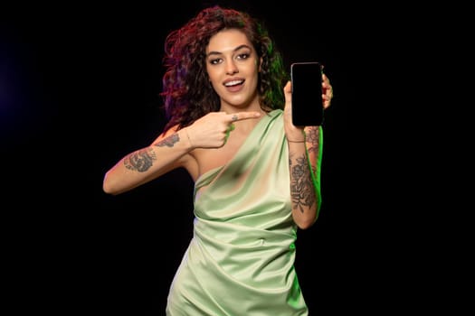 Cheerful excited young brown-haired woman holding smartphone in hand, pointing with finger at blank screen of gadget with copyspace for your lettering. Studio portrait on dark background