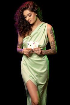 Attractive young female poker player in silk light green dress with long curly hair and tattoos on arms standing on black background, holding pair of winning aces. Gambling concept