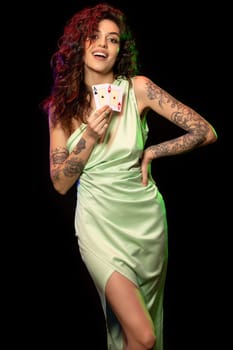 Attractive young poker player girl in silk dress with long curly hair and tattoos on arms standing on black background, playfully showing pair of winning aces. Gambling concept