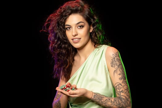 Attractive young dark-haired woman with tattoos on arms holding red and green betting tokens in palms, satisfied with winning poker game. Closeup portrait. Concept of successful gambling