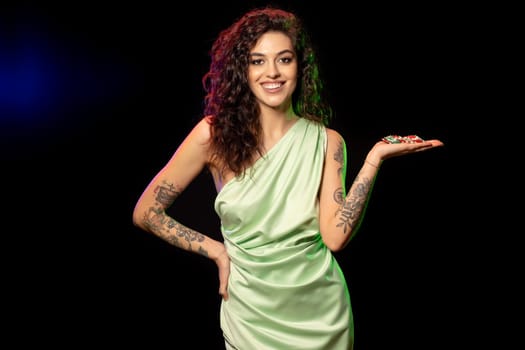 Gambling happy excited young brunette with tattoos on arms standing against black background holding pile of betting chips on palm, cropped shot