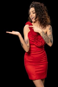 Excited attractive young brunette in red party dress pointing with finger at imaginary product or copyspace for brand lettering on her open palm. Advertising or promotion concept