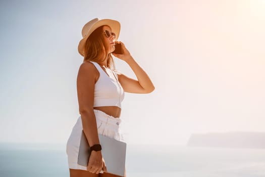 Successful business woman in yellow hat working on laptop by the sea. Pretty lady typing on computer at summer day outdoors. Freelance, travel and holidays concept.