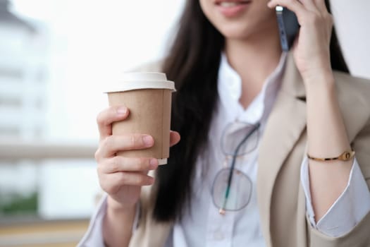 Businesswoman with disposable paper coffee cup talking on mobile phone outside modern office building.
