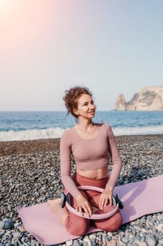 Middle aged well looking woman with black hair doing Pilates with the ring on the yoga mat near the sea on the pebble beach. Female fitness yoga concept. Healthy lifestyle, harmony and meditation.