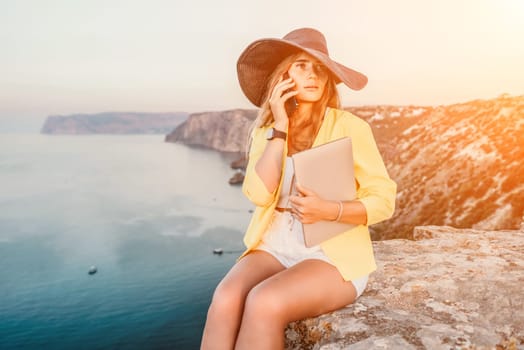 Successful business woman in yellow hat working on laptop by the sea. Pretty lady typing on computer at summer day outdoors. Freelance, travel and holidays concept.