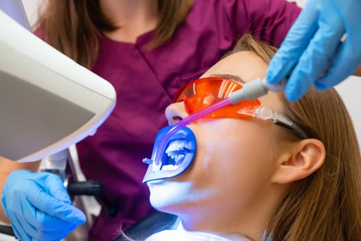 Close up dentist uses saliva ejector while doing a professional whitening of teeth for young patient