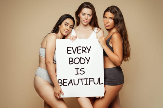 Serious millennial arab, caucasian, latin women in underwear enjoy self love hold banner with inscription every body is beautiful, isolated on beige background, studio. Beauty care, motivation