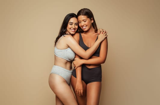 Cheerful millennial latin, arab women in underwear hugs, isolated on beige background, studio. Motivation for self love, spa treatments and fit, wellness, skin care, natural beauty