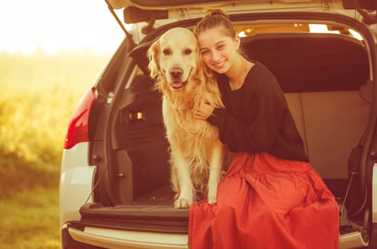 Beautiful girl with golden retriever dog sitting in car trunk and smiling. Pretty teenager hugging purebred pet doggy in vehicle at autumn nature