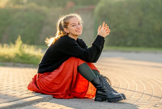 Beautiful teenager girl in red skirt sitting outdoors at street. Pretty teen model posing in trendy clothes