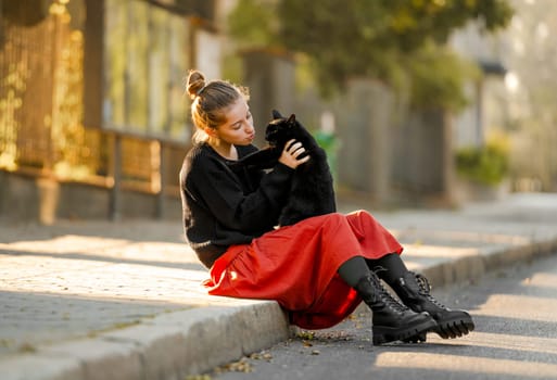 Pretty girl in red skirt hugging black cat outdoors at street with autumn leaves. Beautiful model teenager sitting with feline animal at park