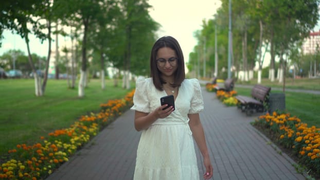 A young woman walks along a flower alley with a phone in her hands. A girl in a white dress looks at her phone and walks among the flowers. 4k