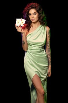 Confident attractive slender young woman with wavy brown hair in elegant light green dress standing against black background, holding pair of aces. Gambling and successful poker game concept