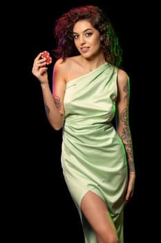 Successful young female poker player in light green dress with naked shoulder and tattoo on arms standing against black background, holding two red betting chips. Gambling concept