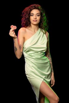 Attractive young brunette with wavy hair and tattoo on arms posing against black studio background, holding pair of betting chips with winning look. Gambling addiction and adult entertainment concept