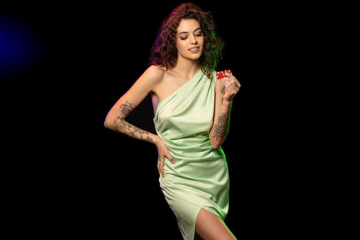 Cheerful stylish young woman with wavy dark hair wearing silk light green dress standing against black background, holding red chips to place bet. Successful gambling concept