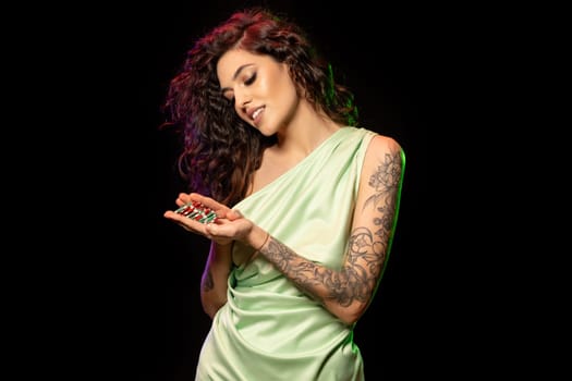 Smiling female poker player standing against black backdrop, contentedly gazing at pile of betting chips in her hand, rejoicing at winning in casino