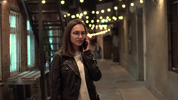 A young woman talks on the phone on a narrow street in the evening. A girl with glasses and a leather jacket on a cozy street. 4k