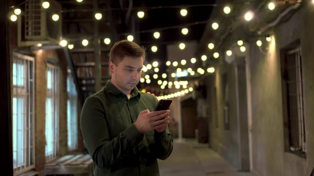 A young man texts on a smartphone on a narrow street in the evening. A man in a shirt on a cozy street with a phone in his hands. 4k