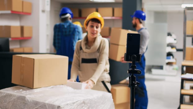 Supervisor recording advertisement video in depot, showing merchandise packages on social media app. Warehouse employee using smartphone to film PR ad, storage room racks.