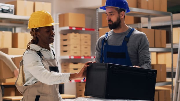 Multiethnic team of people examining list of products on files, talking about merchandise stock for inventory. Man and woman in overalls checking goods in storage room depot, industrial work.