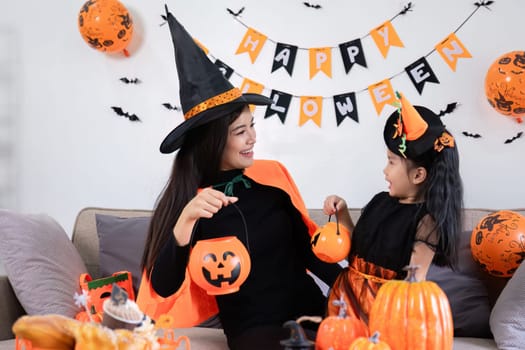 A playful young mother sits on a comfortable sofa in the living room decorated with pumpkins and her cute little daughter tells a spooky Halloween story. Funny mother and girl having fun at home.