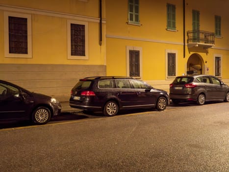 Cremona, Lombardy, Italy October 15th 2024 Cars parked on the side of a street at night, illuminating the facade of a yellow building with light reflecting on the wet asphalt