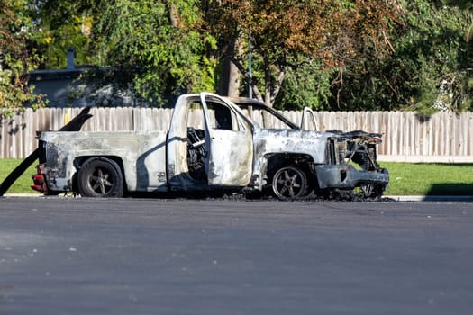 A burnt-out truck sits abandoned on a suburban street, its charred frame highlighting recent fire damage. Bright sunlight and greenery contrast with the vehicle's destruction, adding to the scene's stark realism.